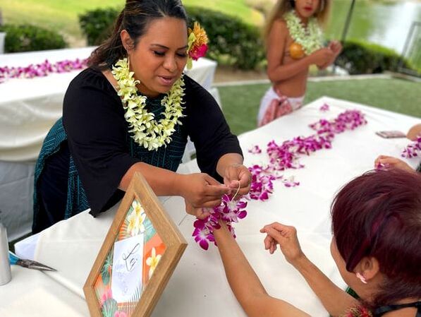 A lei maker, adorned with a double lime ginger lei, is teaching a woman how to craft an orchid lei bracelet. The focus is on their hands as they intricately weave the orchids, highlighting the traditional art of lei making.