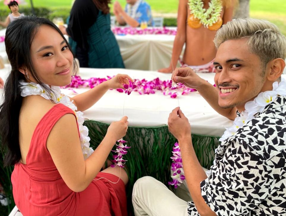A Hawaiian couple engaged in the traditional art of lei making, working side by side to create beautiful orchid leis. The image captures their shared focus and the intricate details of the delicate orchids.d