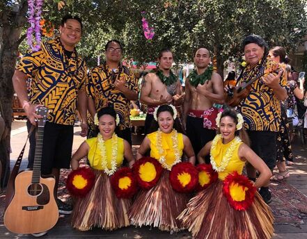 Female and male Hawaiian hula dancers posing with a live band, all wearing Samoan-designed shirts, also known as island button-ups. The scene captures a harmonious blend of dance and music, showcasing the vibrant cultural attire and the lively atmosphere of the event.