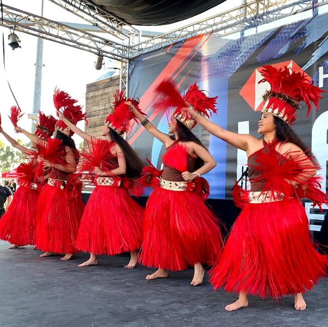 Tahitian dancers performing a traditional dance, dressed in colorful, modest Tahitian attire, featuring vibrant sarongs and floral headdresses, set against a backdrop of a festival stage