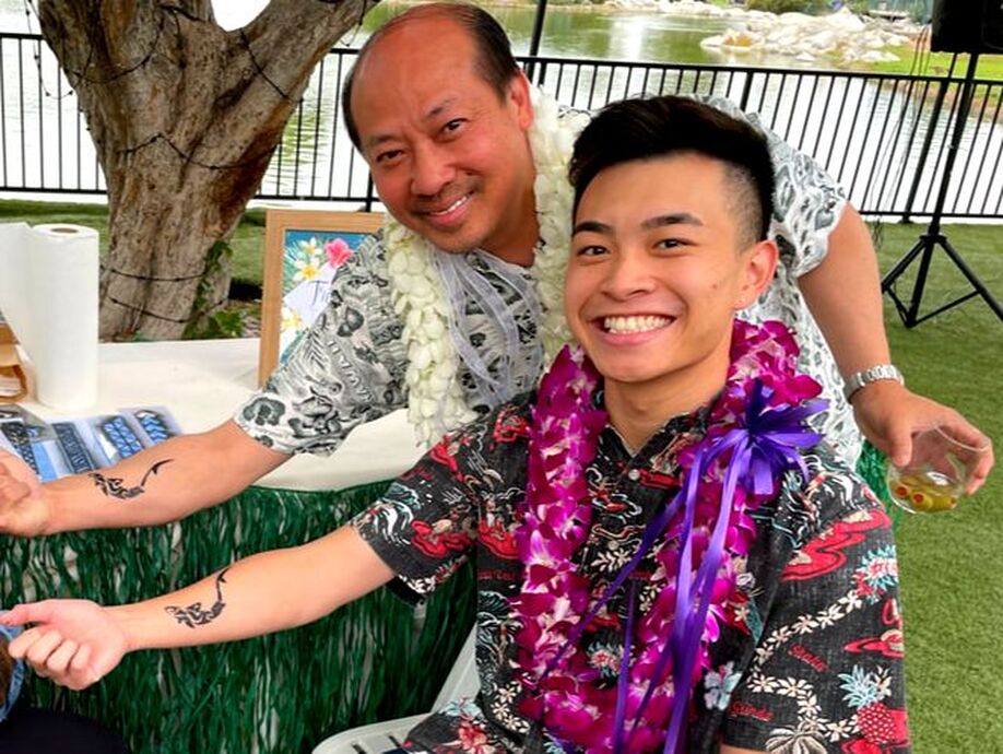 Father and son in fresh orchid leis enjoy airbrushed polynesian and Maori tattoos on a beautiful venue with a lake