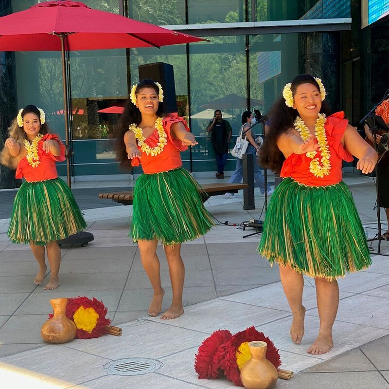 Three hula dancers performing outdoors at a corporate luau in Southern California, gracefully using traditional hula implements. They are set against a backdrop of a professionally arranged outdoor event space.