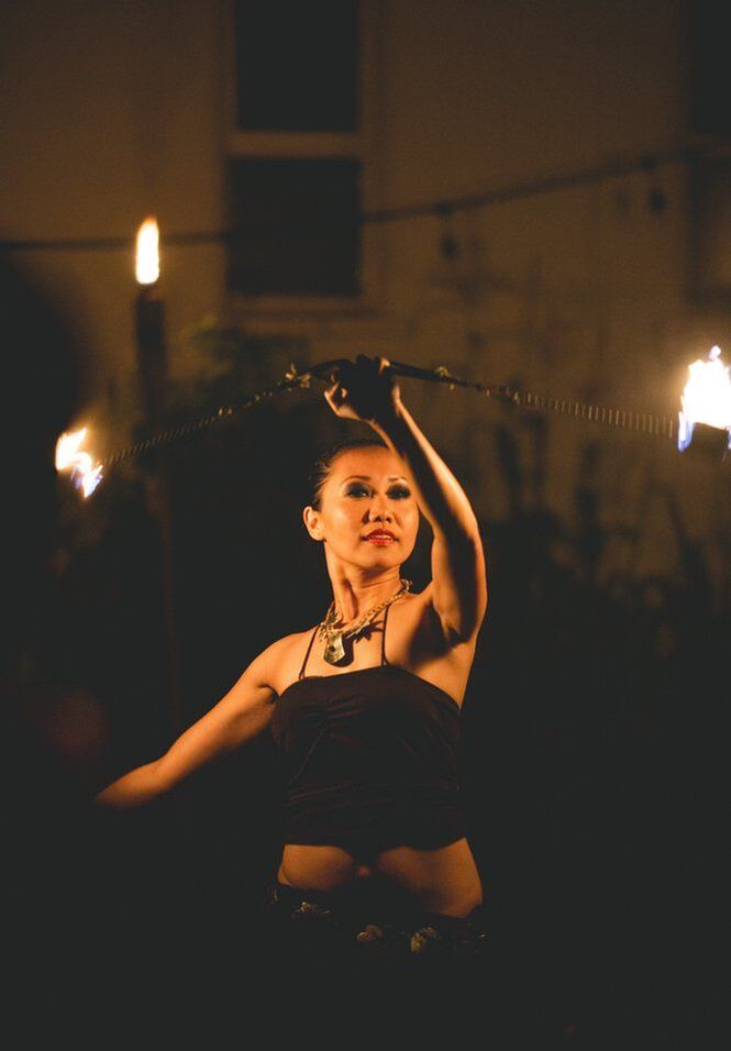 Fire poi dancers performing in a dimly lit venue, their movements both alluring and dangerous yet graceful. The flames create captivating patterns in the air, highlighting the skill and artistry of the dance.