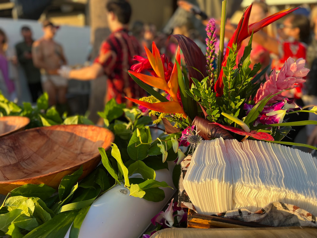 Island-style catering set up in Southern California on the beach. Tropical floral centerpieces, greenery covered buffet, and wooden serve ware to fit an island-style catering event.